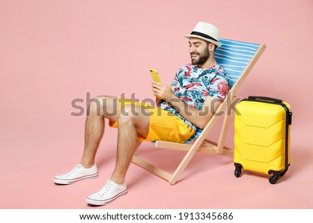 Full length smiling young traveler tourist man in hat sit on deck chair using mobile cell phone typing sms message isolated on pink background. Passenger travel on weekend. Air flight journey concept