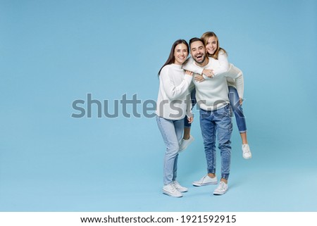 Full length of funny young parents mom dad with child kid daughter teen girl in sweaters giving piggyback ride to joyful, sitting on back isolated on blue background. Family day parenthood concept