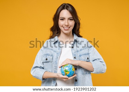 Young smiling happy brunette fun geography student woman 20s wearing stylish casual denim shirt white t-shirt holding in palms Earth world globe isolated on yellow color background studio portrait.