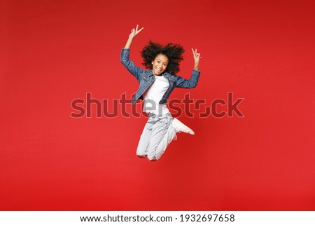 Full length of funny little african american kid girl 12-13 years old in denim jacket jumping showing victory sign isolated on red background children studio portrait. Childhood lifestyle concept