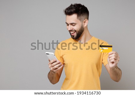 Young caucasian smiling bearded attractive rich student man 20s wear casual yellow basic t-shirt hold mobile cell phone credit bank card shopping online isolated on grey background studio portrait