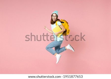 Full length side view of excited young woman student in blue t-shirt hat glasses backpack hold notebooks jump isolated on pink background studio. Education in high school university college concept