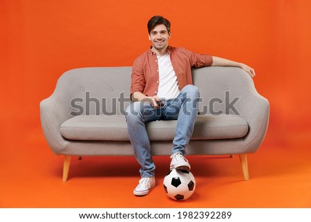 Young man football fan in shirt support favorite team with soccer ball sit on sofa at home watch tv live stream switch channel isolated on orange background. People sport leisure lifestyle concept.
