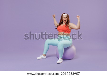 Full length side view young chubby overweight plus size big fat fit woman in red top warm up train sit on fit ball do winner gesture isolated on purple background gym. Workout sport motivation concept