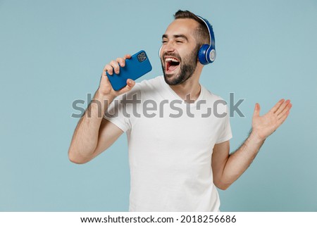 Young man wear blank print design white t-shirt headphones listen to music sing song record voice by mobie cell phone dictaphone isolated on plain pastel light blue color background studio portrait.