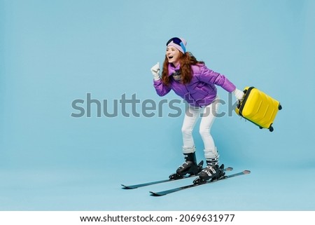 Full body traveler tourist woman in winter ski windbreaker jacket goggles hold valise suitacase isolated on plain blue background studio Passenger travel abroad weekends getaway Air flight concept.