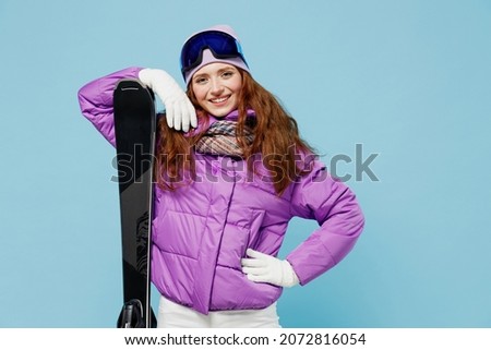 Skier cheerful satisfied happy sporty woman wear warm purple padded windbreaker jacket goggles mask spend extreme weekend in mountains hold ski stand akimbo isolated on plain blue background studio.