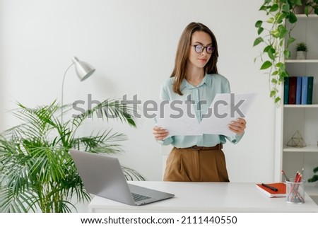Young successful employee business woman in blue shirt hold paper account documents work stand at workplace white desk with laptop pc computer at light modern office indoors Achievement career concept Foto stock © 