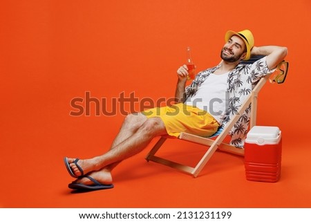 Young smiling happy fun cool tourist man in beach shirt hat hold beer bottle lie on deckchair near fridge isolated on plain orange background studio portrait. Summer vacation sea rest sun tan concept Foto stock © 