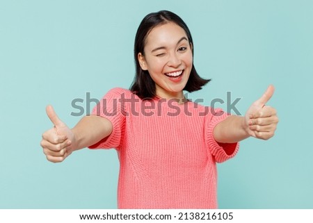 Young smiling fun happy woman of Asian ethnicity 20s wearing pink sweater showing thumb up like gesture blink isolated on pastel plain light blue background studio portrait. People lifestyle concept Foto stock © 