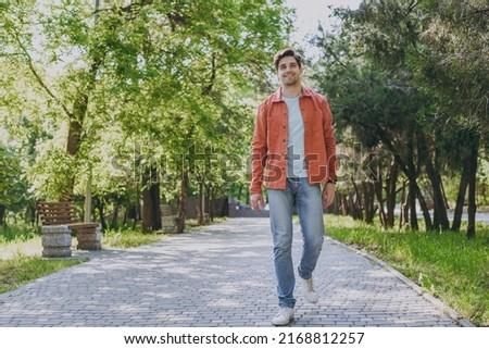 Full length young man 20s he wearing orange jacket blue t-shirt walking look camera rest relax in spring green city park go down alley sunshine lawn outdoors on nature. Urban lifestyle leisure concept ストックフォト © 