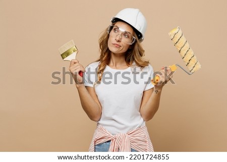 Young minded employee laborer handyman woman wear white t-shirt helmet choosing roller brush paint walls isolated on plain beige background Instruments accessories for renovation room Repair concept Foto stock © 