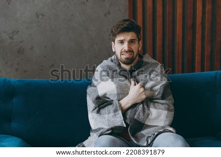 Young sick ill unhappy displeased cold man wear red t-shirt wrapped in plaid shivering sit on blue sofa couch stay at home hotel flat spend time in living room indoors grey wall. People health concept Foto stock © 