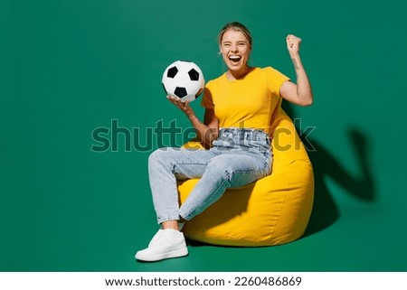 Full body young fun woman fan wear basic yellow t-shirt cheer up support football sport team sit in bag chair hold soccer ball watch tv live stream do winner gesture isolated on dark green background Foto stock © 