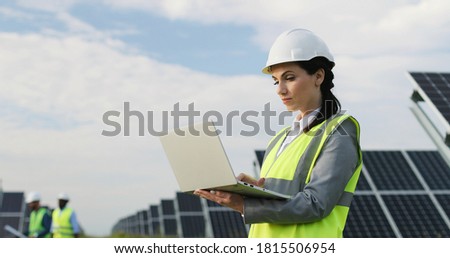 Beautiful business lady standing among field of solar panels in special uniform. Female engineer uses laptop to work outside.