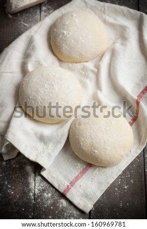 Three floured balls of uncooked homemade pizza dough proofing on a kitchen towel on a rustic dark wooden table, taken from above.