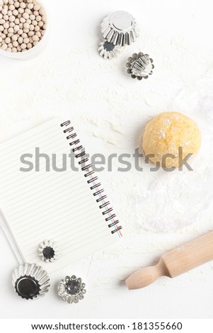 Unrolled and unbaked shortcrust pastry dough with assorted baking tools: rolling pin, ceramic baking beans, mini tart tins and blank recipe book. Taken on a floured white surface, directly from above.