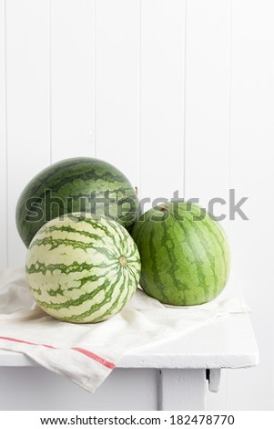 Three different watermelons on a kitchen towel and placed on a white vintage farmhouse table. Taken against a white wood plank wall. Styled in a rustic cottage manner.