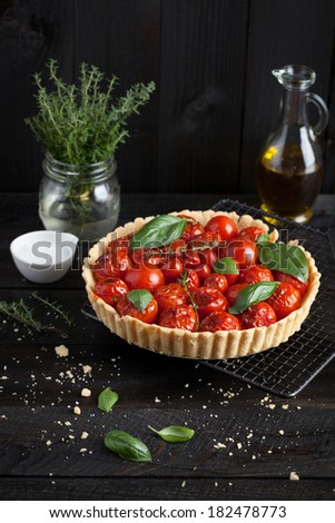 Cherry tomato tart with fresh basil leaves and thyme served on a cooling rack. Placed on a black table with a bottle of olive oil, sea salt and a fresh thyme. Taken against a black wood plank wall.