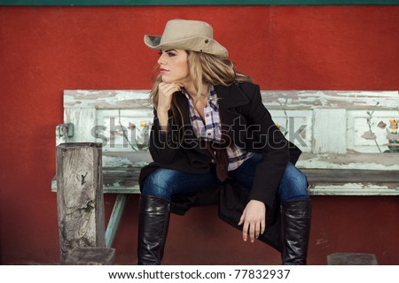 Country style fashion portrait of a beautiful long-haired blond young woman wearing a cowboy hat and sitting on an old bench in front of a red wall. Shallow depth of field.