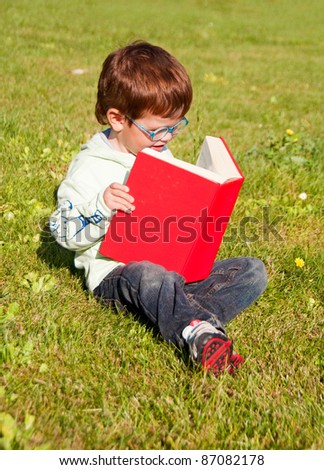 Child with glasses reading a red book sitting on grass