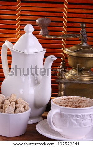 A grouping comprising of a coffee pot, sugar bowl containing brown sugar cubes, cup of coffee and a coffee bean grinder, bamboo background.