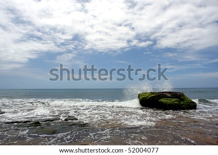 Seascape showing a sea sprayed large green rock as the tide comes in the sky is blue with white clouds