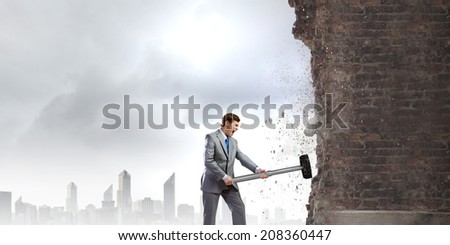Young determined businessman with big hammer in hands crashing wall
