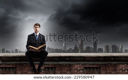 Young businessman sitting on building top with book