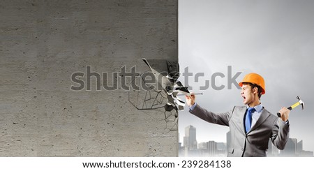 Young businessman in suit and hardhat hammering nail in wall