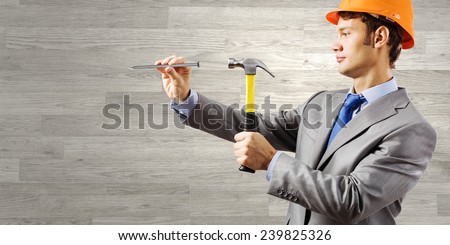 Young businessman in suit and hardhat hammering nail in wall