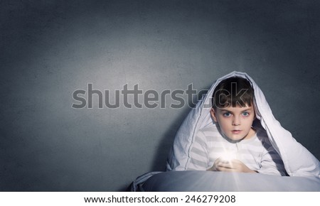 Little cute boy sitting in bed under blanket with flashlight