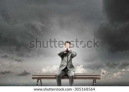 Young shocked businessman sitting on wooden bench and covering eyes with palms