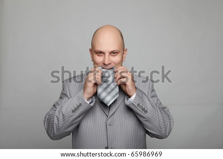 Bald businessman in a gray suit with a gray background eats his own tie. Funny business