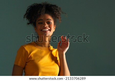 Beautiful happy dark skinned african american ethnicity woman in yellow tshirt raising hand in greeting while looking in camera with pleasant smile, posing against gray green studio wall background 商業照片 © 