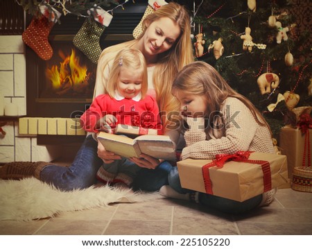 Young mother and her two little daughters with Christmas gifts reading the book by a Christmas tree in cozy living room with fireplace in winter
