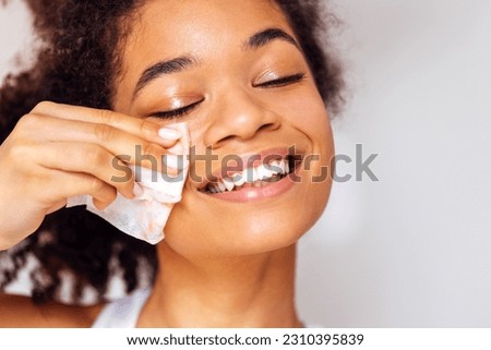 Close up of face of young happy attractive african washing off her makeup. Charming smiling mixed race girl wipes her face skin with napkin. Young beautiful woman closed her eyes. Skin care concept. Foto stock © 
