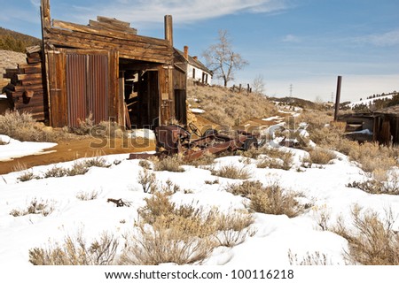 A garage that once served a deserted mining community slowly disintegrates and falls apart. A house is in the background and the chassis of an old automobile across the road is in the foreground
