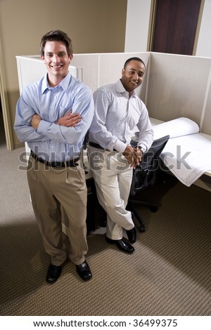 Two male colleagues standing in cubicle with blueprints