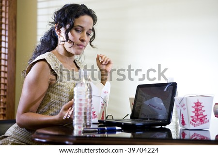 Female Hispanic office worker eating Chinese takeout food for lunch while working on laptop