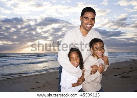 Happy African-American father,  ten year old son and six year old daughter hugging on beach
