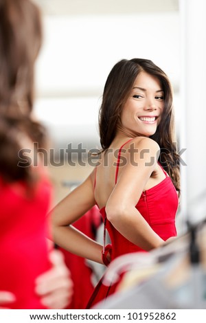 Woman trying clothes / red dress in clothing store changing room looking at mirror smiling happy. Beautiful young multiethnic girl in clothing store.