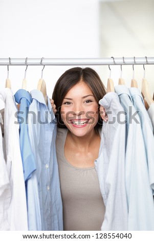 Small business clothing shop owner portrait in store. Funny image of woman clothes shop owner peeping through shirts smiling happy and excited at camera. Multicultural Asian Caucasian female model.