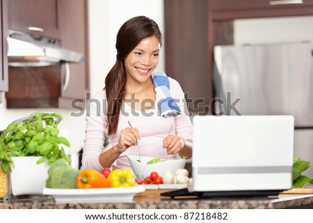 Cooking woman looking at computer while preparing food in kitchen. Beautiful young multiracial woman reading cooking recipe or watching show while making salad.