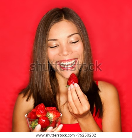 Strawberries - woman eating strawberry smiling happy on red background. Beautiful healthy eating mixed race Chinese Asian / Caucasian girl joyful inside.