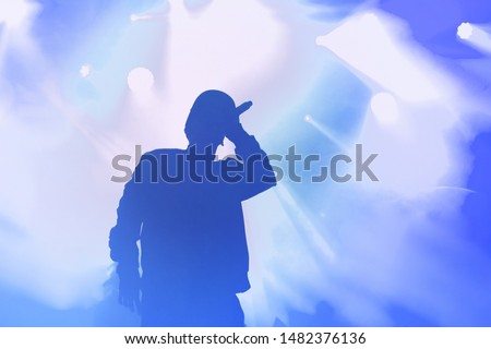 Stock photo of young rap singer with mic in hand singing popular song on stage in blue lights.Hip hop artist performing live on scene in music hall.Repper with microphone in royalty free backgrounds