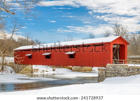 The red Cataract Falls Covered Bridge in rural Indiana crosses Mill Creek in a snowy winter landscape with a cloudy blue sky.