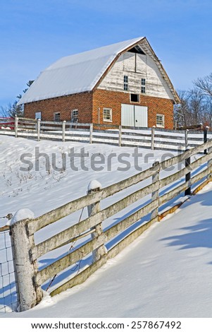 An old red brick barn and a rugged white fence are covered in winter snow on a sunny day.