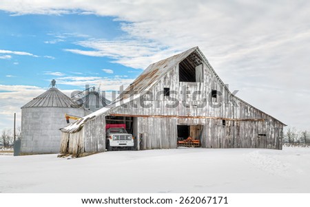 A snowy winter landscape surrounds a large, weathered white barn and farm equipment.