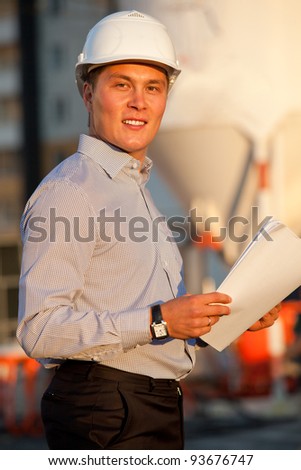 portrait of well-dressed man with documants in hard hat standing against the construction